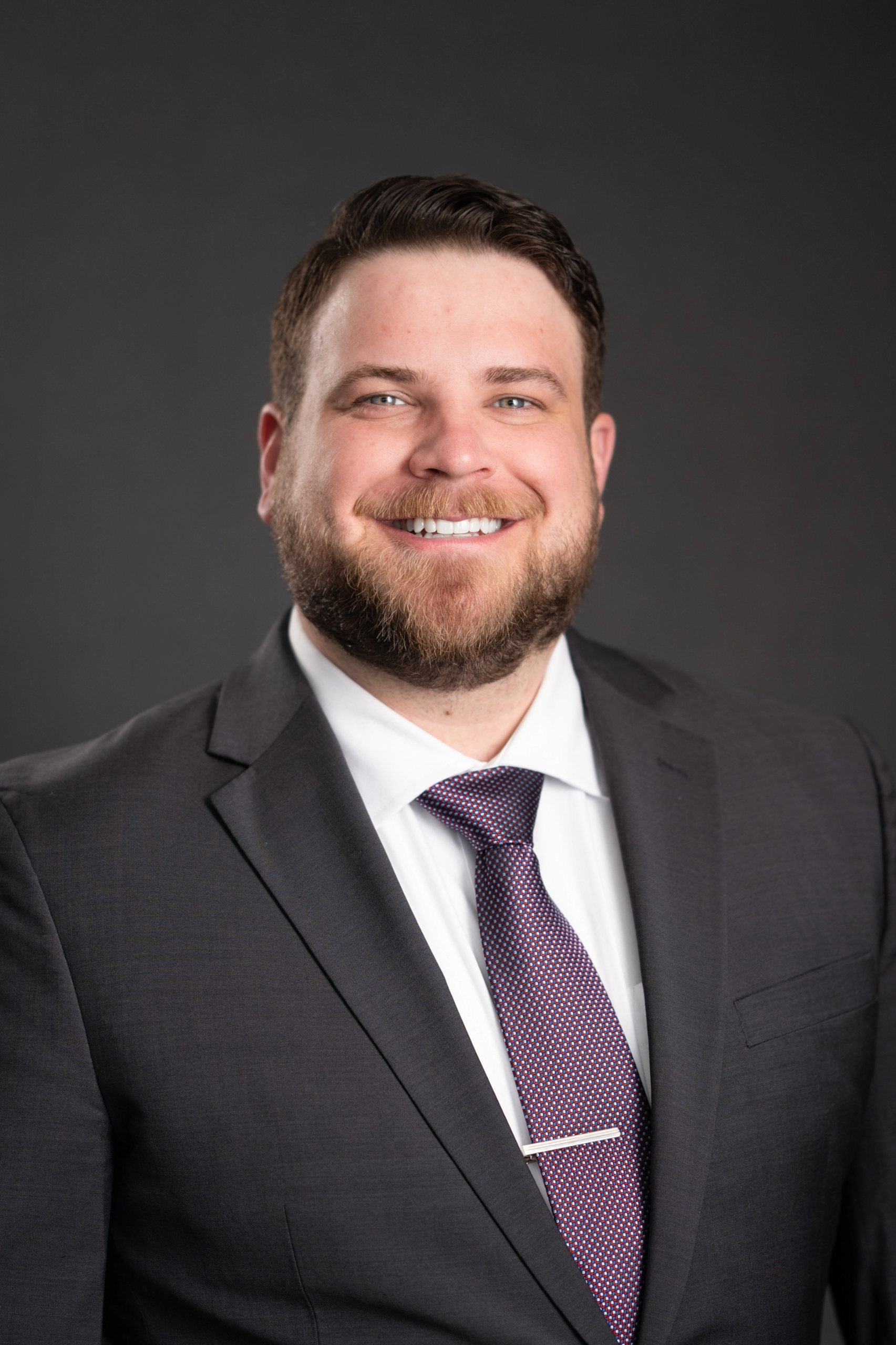Head-and-Shoulders portrait of a smiling man in a dark suit against a gray background.